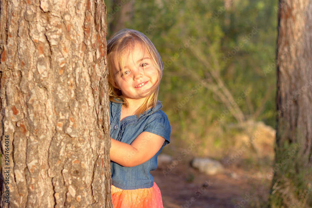 A cute blonde little girl playing hide and seek behind a tree at sunset A cute blonde little girl playing hide and seek behind a tree at sunset