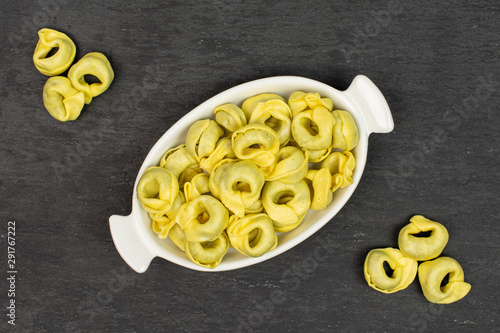 Lot of whole fresh yellow spinach filled tortelloni in white oval ceramic bowl flatlay on grey stone