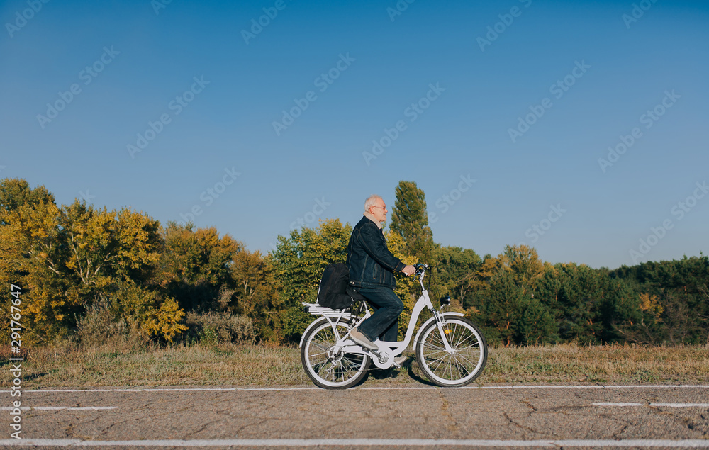 Obraz premium An elderly man, a pensioner rides an electric white bike on an asphalt road against the background of autumn nature. The concept of a happy old age.