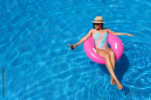 Young woman with sunglasses, hat and swimsuit in a blue pool. Pretty girl on a pink float enjoying the summer while having a cocktail.