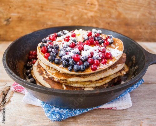 Stack of homemade shrovetide carnival clean eating oat pancakes with powered sugar, blueberries and cowberries in cast iron pan on rustic country table. Soft focus. Copy space