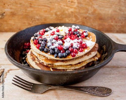 Stack of homemade shrovetide carnival clean eating oat pancakes with powered sugar, blueberries and cowberries in cast iron pan on rustic country table with old silver dishware. Soft focus. Copy space