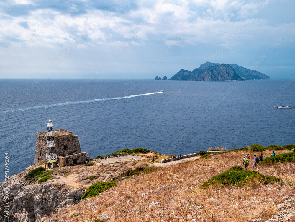 Hikers walk in Punta Campanella with the lighthouse, the Tower of Minerva and the island of Capri with the faraglioni in the background. Protected marine natural area of Punta Campanella. Sorrento Pen