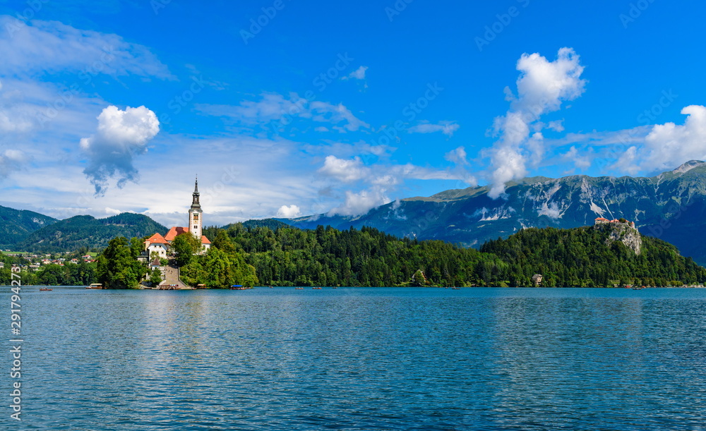 The scenery of Lake Bled with a medieval castle, an island with a church and alpine mountains in the background. Bizarre vertical clouds.