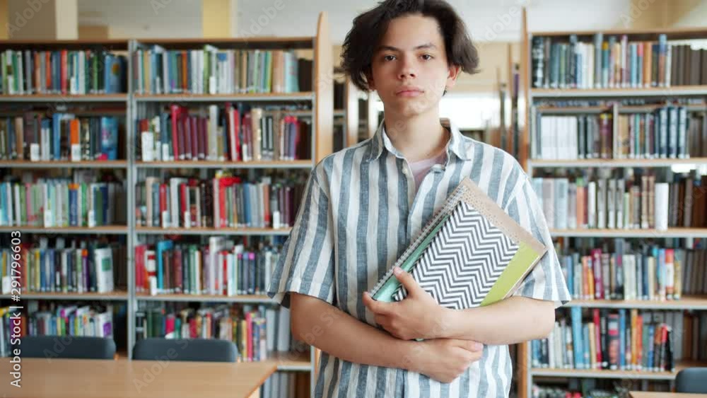 Slow motion portrait of good-looking teenage student standing in library with books and looking at camera with serious face. People and knowledge concept.