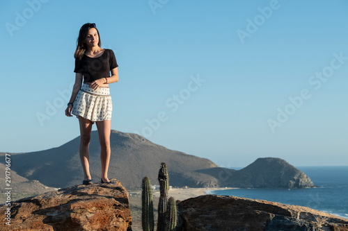 girl standing in a rock with hills and a beach on the background with the sunset