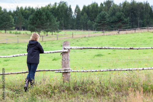 Woman enjoying rural view, standing her back to camera, keeping hands on wooden fence