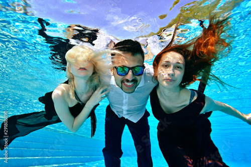 A group of young people swim and pose for the camera underwater in the pool in fashionable dresses. Girls, blonde and red, hugging a guy in sunglasses. The guy smiles at the camera. Fashion portrait