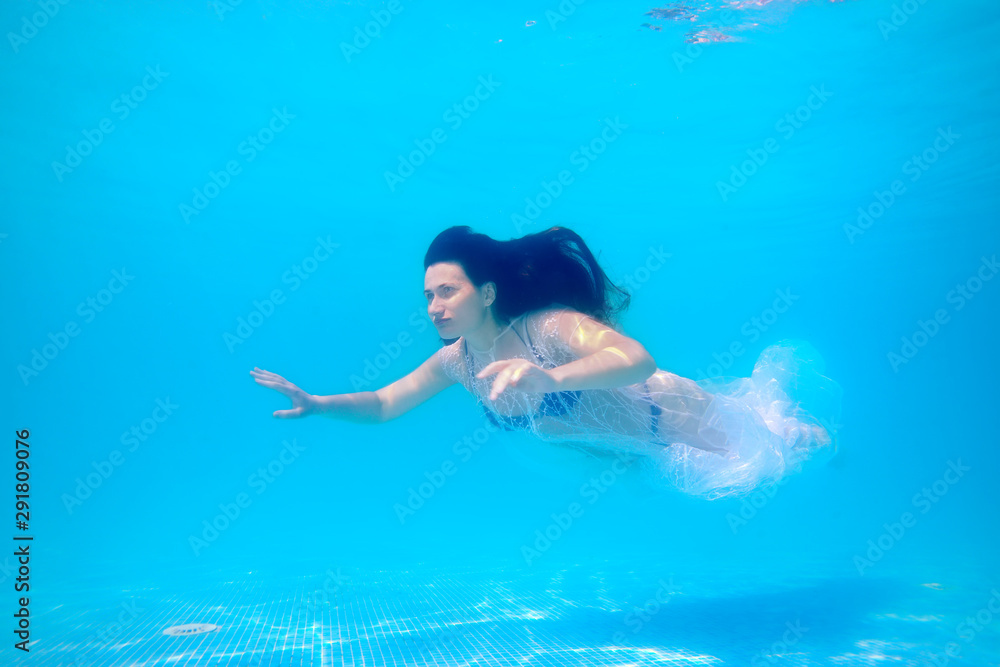 A beautiful sporty girl is swimming underwater in a blue swimsuit and a white transparent Cape, with long loose hair on a bright Sunny day on a blue background. Concept