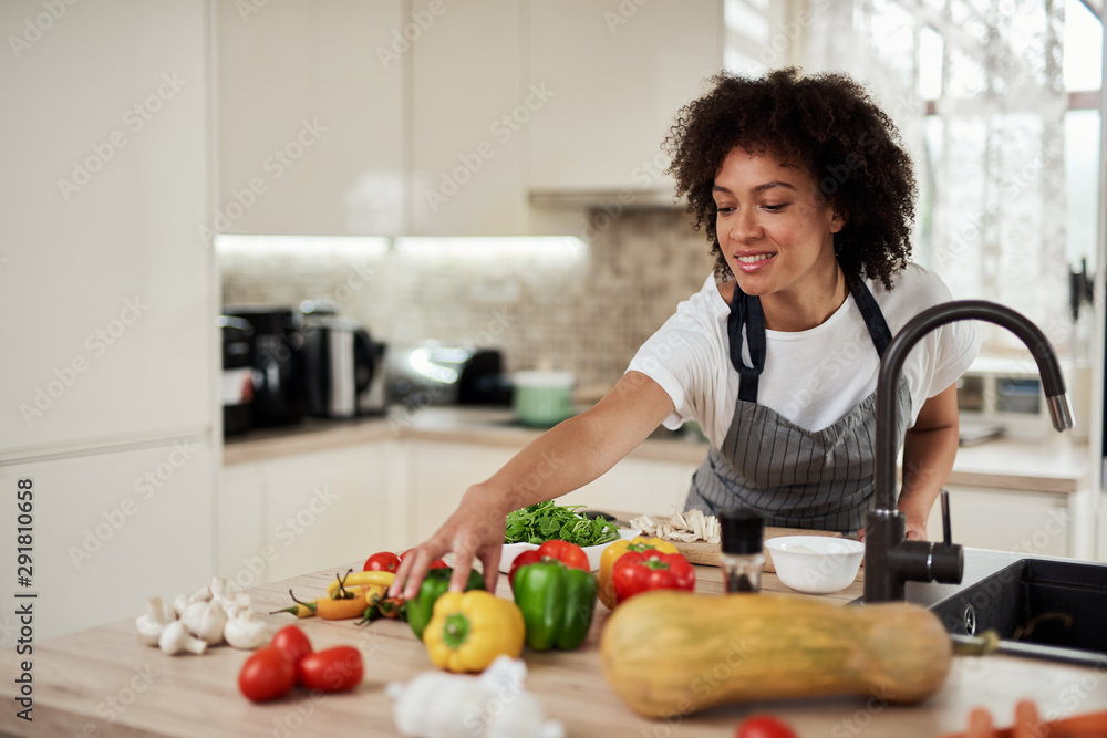Beautiful mixed race housewife reaching for green pepper while standing in kitchen. Dinner preparation.