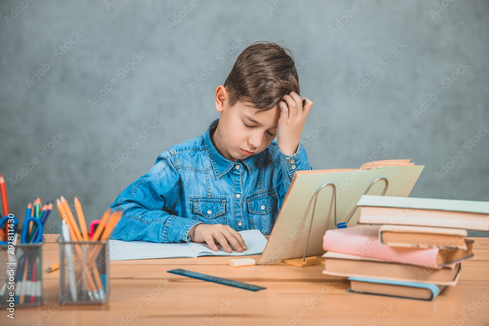 Little tired schoolboy is sitting at the desk, overwhelmed with books, holding hand on the forehead. Little tired schoolboy is sitting at the desk, overwhelmed with books, holding hand on the forehead.