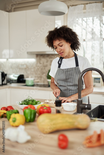 Gorgeous mixed race housewife in apron standing in kitchen and chopping mushrooms. On kitchen counter are all sorts of vegetables.