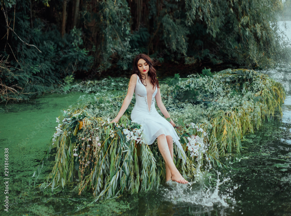 Fototapeta premium Cheerful woman in a white vintage outfit sits on a boat and splashes her bare feet in the water. wild background river with water lilies and green trees. Happy Feminine Princess deep neckline dress