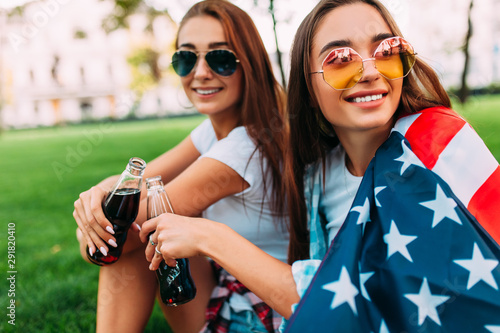 Two attractive young girls in sunglasses sitting in the Park on the lawn with the American flag, with a refreshing drink