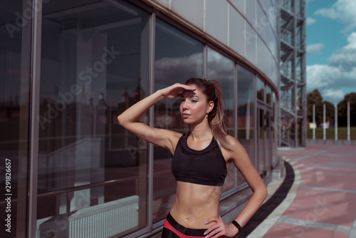 Beautiful athletic tanned girl looks into distance  covers her hand from sun  in summer city background of glass windows  free space for fitness motivation text. Sportswear top.