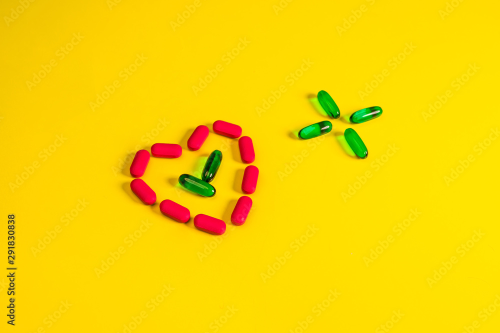 Fototapeta premium Green transparent capsules folded in the form of a cross next to a heart from capsules on a yellow background.The concept of pharmacology.