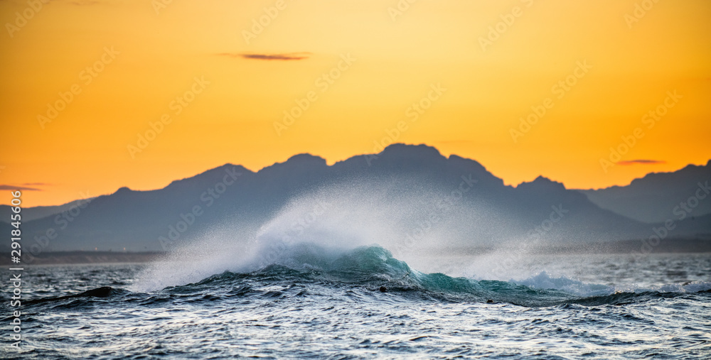 Seascape.  Red dawn sky, waves crashing with splashes against stones, silhouettes of mountains on the horizon.  South Africa.