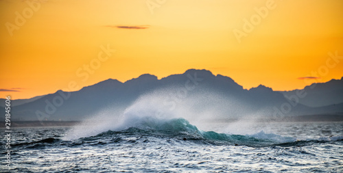 Seascape. Red dawn sky, waves crashing with splashes against stones, silhouettes of mountains on the horizon. South Africa.