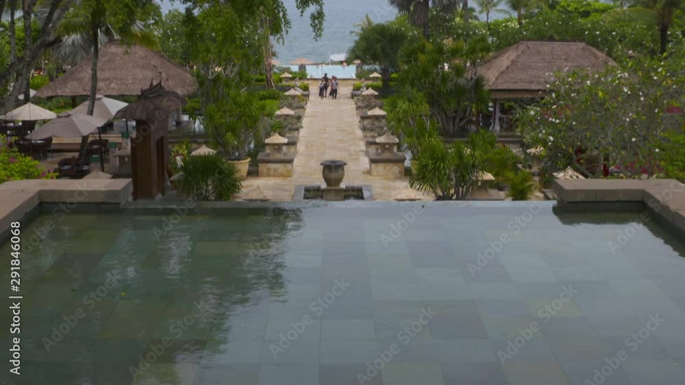 Wide low angle tilting shot of clear pool water with patterned floor tiles , resort buildings surrounded by lush green trees, and walking people approaching through a pathway from the beach, coastal Wide low angle tilting shot of clear pool water with patterned floor tiles , resort buildings surrounded by lush green trees, and walking people approaching through a pathway from the beach, coastal