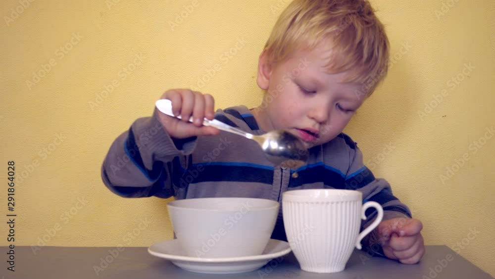 Small blond child sits at table with two cups on yellow background. Boy pours hot tea from one cup into another with spoon. Baby comes up with entertainment and is actively learning necessary skills Small blond child sits at table with two cups on yellow background. Boy pours hot tea from one cup into another with spoon. Baby comes up with entertainment and is actively learning necessary skills