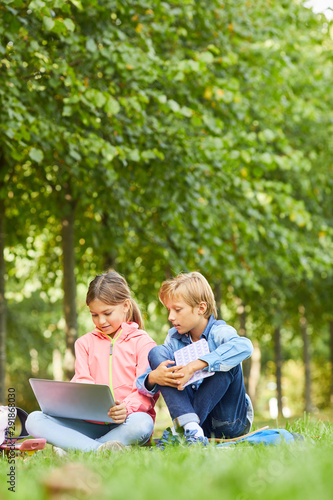 Girl using laptop computer together with her classmate while they sitting on green grass outdoors