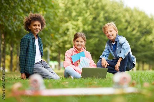 Portrait of happy school chidlren sitting together on the grass and using laptop and book in their study outdoors photo