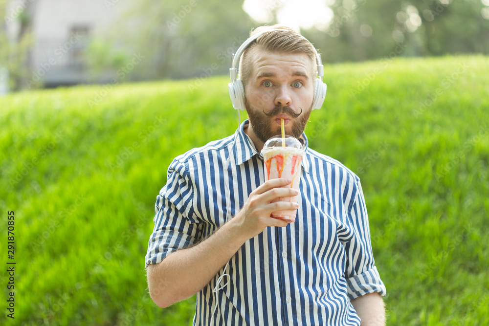 Fototapeta premium Side view of a stylish young man with a beard holding a milkshake and admiring the city views walking in the park on a warm summer day. The concept of rest and relaxation.