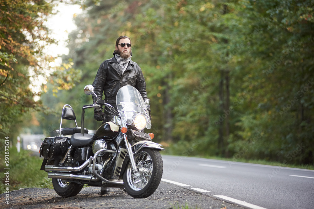 Bearded tall motorcyclist in dark sunglasses, black leather jacket standing at shiny modern powerful cruiser motorbike on blurred background of hilly asphalt road and green trees.