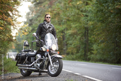 Bearded tall motorcyclist in dark sunglasses  black leather jacket standing at shiny modern powerful cruiser motorbike on blurred background of hilly asphalt road and green trees.