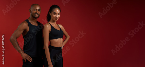 Training together. Young and cheerful african fitness couple in sportswear looking at camera with smile while standing against red background