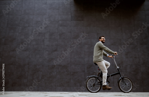 Full length of handsome fashionable caucasian man in sweater and with eyeglasses sitting on the bike. In background is gray wall.
