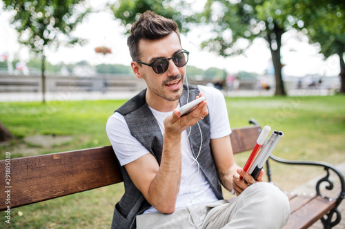 Papier peint Young blind man with smartphone sitting on bench in park in city, calling