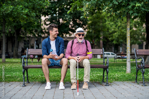 Young man and blind senior with white cane sitting on bench in park in city. photo