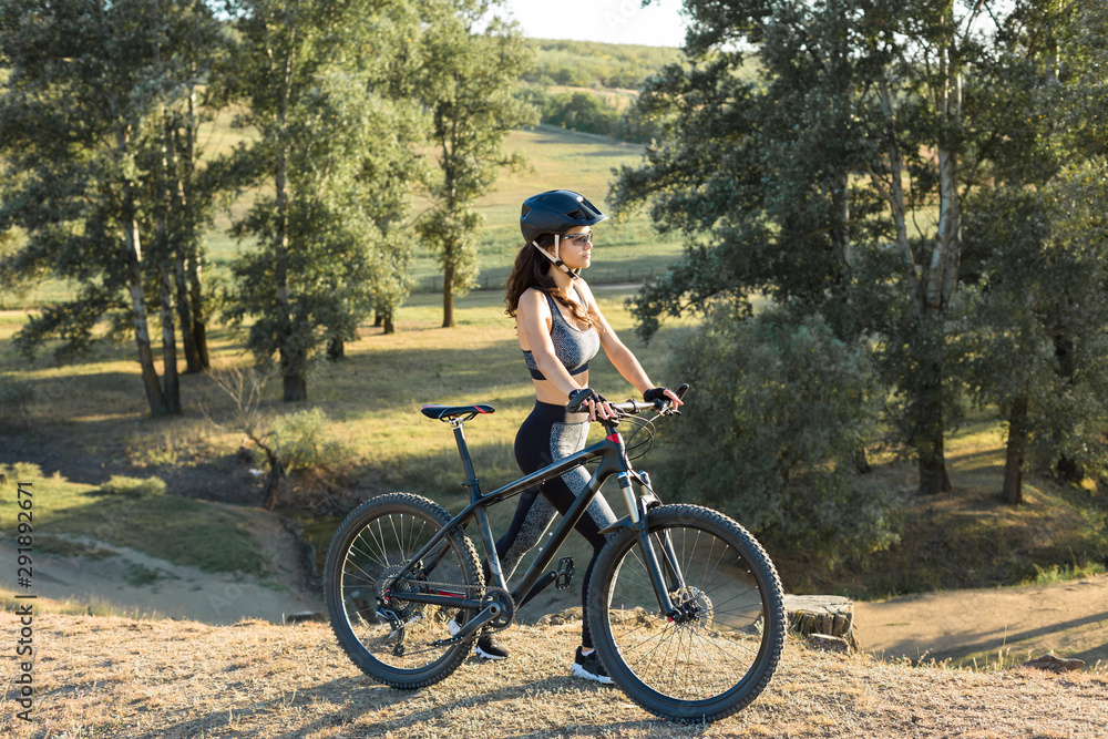 Fototapeta premium Girl on a mountain bike on offroad, beautiful portrait of a cyclist at sunset, Fitness girl rides a modern carbon fiber mountain bike in sportswear, a helmet, glasses and gloves. 
