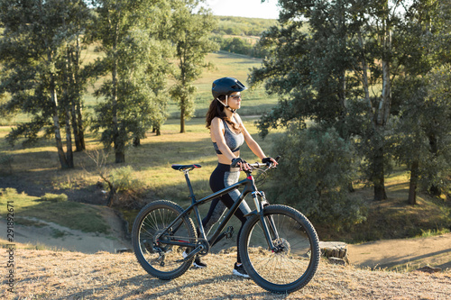 Girl on a mountain bike on offroad, beautiful portrait of a cyclist at sunset, Fitness girl rides a modern carbon fiber mountain bike in sportswear, a helmet, glasses and gloves.	