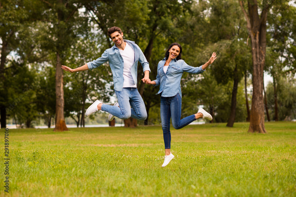 Full length photo of cute pair holding hands jumping high in green park wear denim outfit Full length photo of cute pair holding hands jumping high in green park wear denim outfit