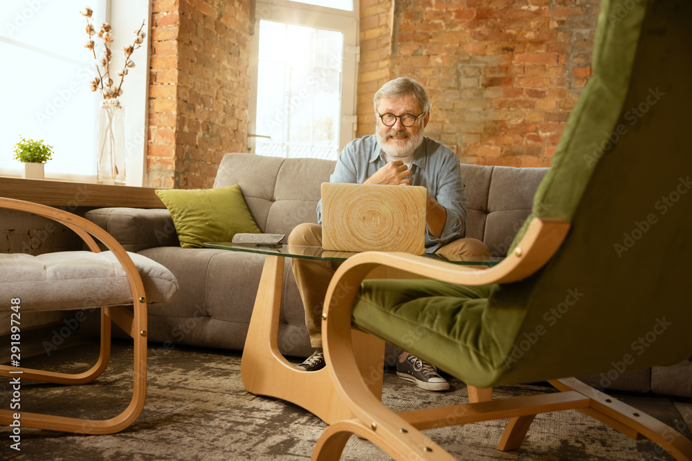 Fototapeta premium Senior man working with laptop at home - concept of home studying. Caucasian male model sitting on sofa and doing his homework or serfing in internet, watching cinema or webinar, playing games.