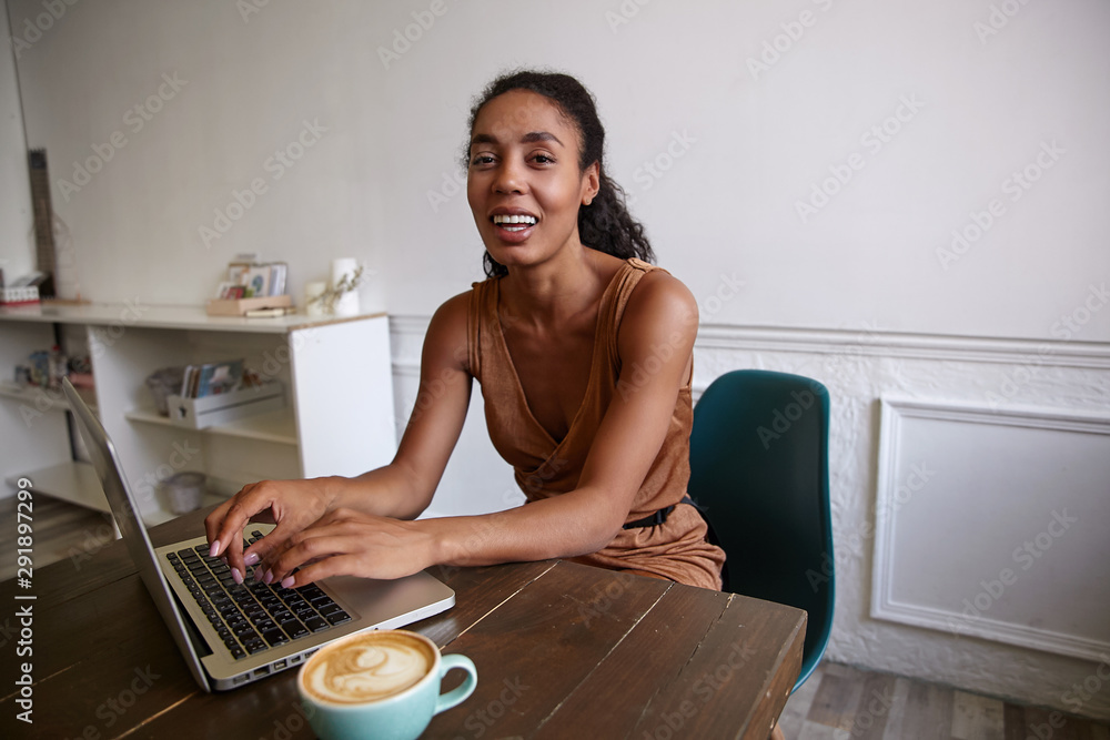 Positive dark skinned woman with curly hair working remotely in coffee house, keeping her hands on keybourd and looking to camera joyfully Positive dark skinned woman with curly hair working remotely in coffee house, keeping her hands on keybourd and looking to camera joyfully