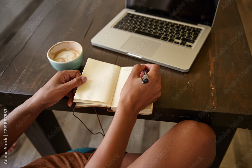 Indoor side view of female hands with pen and notebook, writing notes while having cup of coffee, using modern laptop for remote work