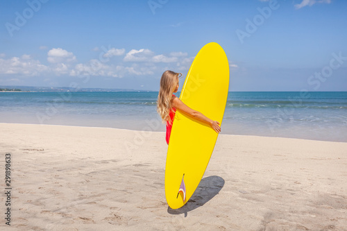 Back view of young woman with long hair in red swimwear holding the bright yellow surfboard on beach with sea and blue sky on background on Bali island, Indonesia. Active lifestyle. Holiday leisure.