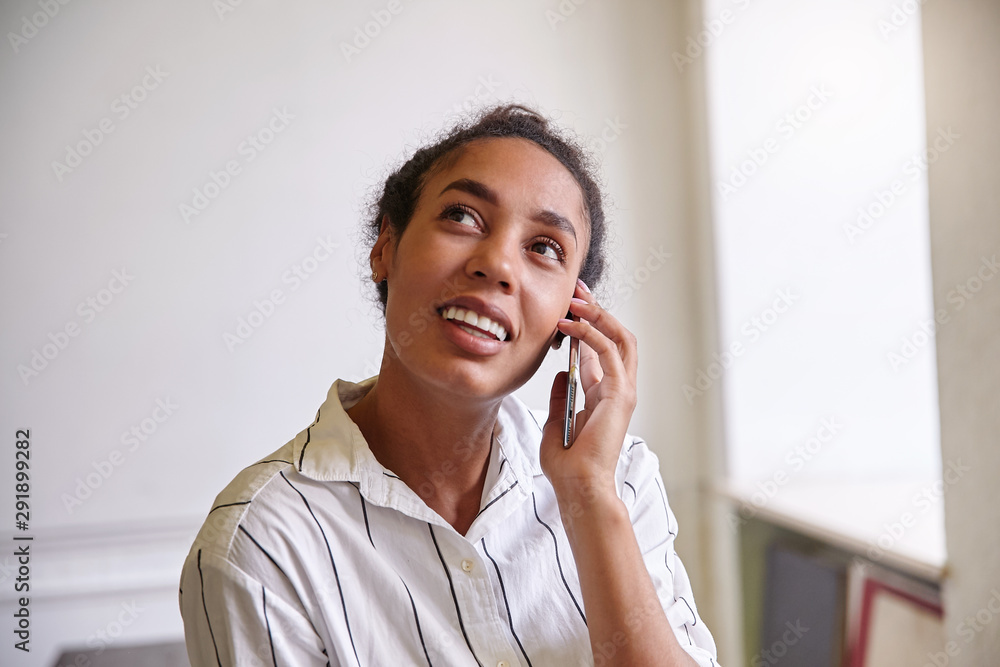Close-up of charming dark skinned woman with curly hair giving call with her mobile phone, looking upwards and smiling cheerfully, wearing casual shirt Close-up of charming dark skinned woman with curly hair giving call with her mobile phone, looking upwards and smiling cheerfully, wearing casual shirt