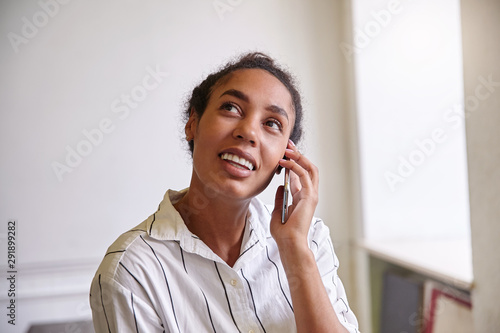 Close-up of charming dark skinned woman with curly hair giving call with her mobile phone, looking upwards and smiling cheerfully, wearing casual shirt