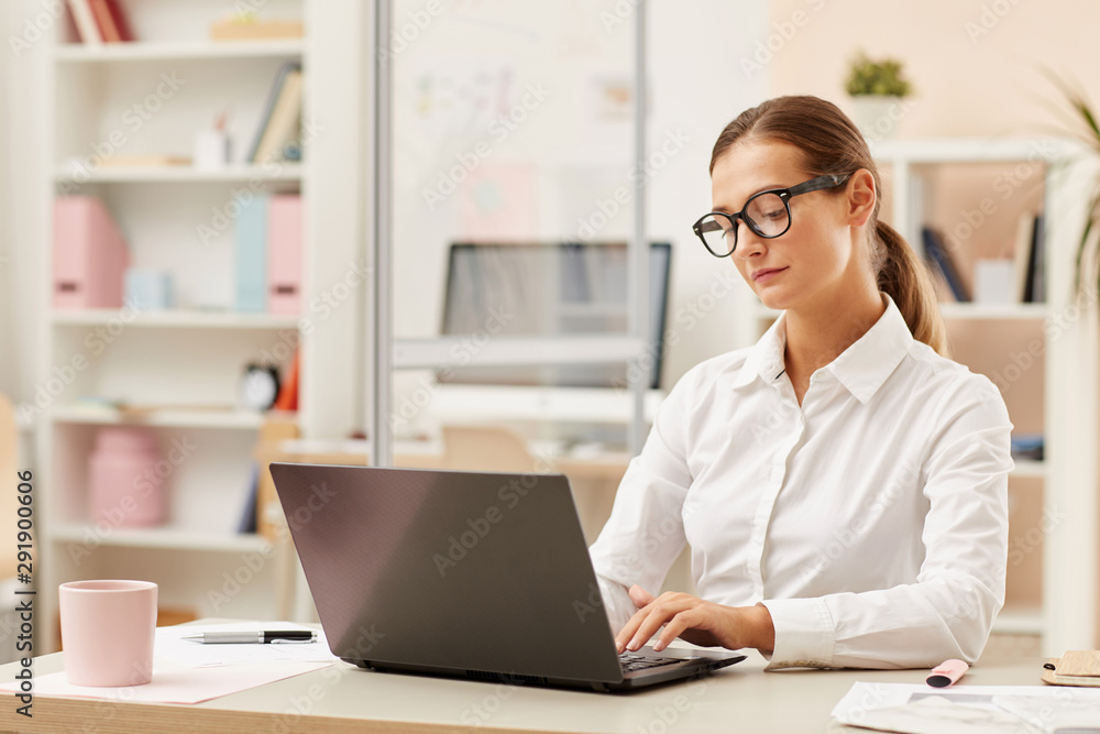 Young secretary in eyeglasses and in white shirt sitting at the table and working online on laptop at office Young secretary in eyeglasses and in white shirt sitting at the table and working online on laptop at office