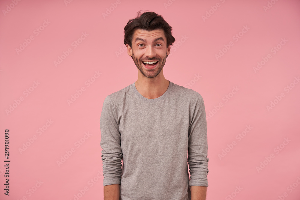 Indoor portrait of happy bearded male with trendy haircut wearing casual clothes, posing over pink background with hands down, looking at camera cheerfully with wide mouth opened Indoor portrait of happy bearded male with trendy haircut wearing casual clothes, posing over pink background with hands down, looking at camera cheerfully with wide mouth opened