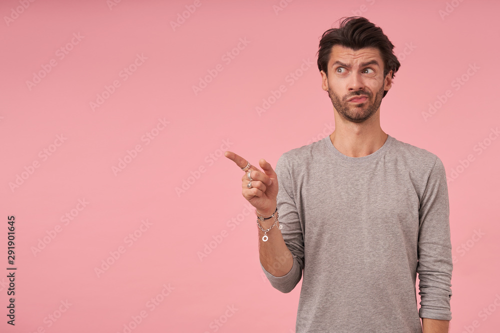 Studio photo of dark haired man with beard standing over pink background, pointing aside with forefinger, looking at camera with doubting face and pursing lips