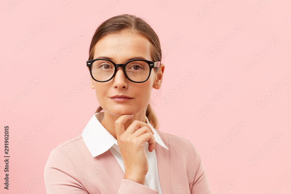Portrait of young businesswoman in eyeglasses and in pink suit looking at camera with serious expression isolated on pink background Portrait of young businesswoman in eyeglasses and in pink suit looking at camera with serious expression isolated on pink background