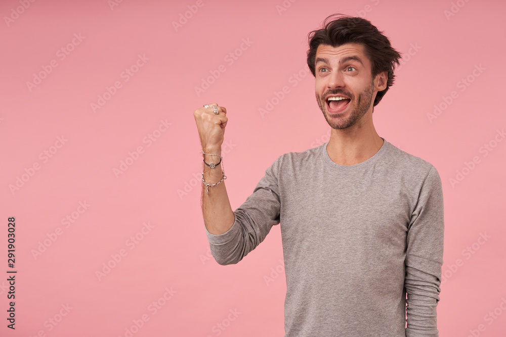 Happy handsome male with trendy haircut wearing grey sweater, standing over pink background, looking aside and raising fist in yes gesture, being happy and smiling widely
