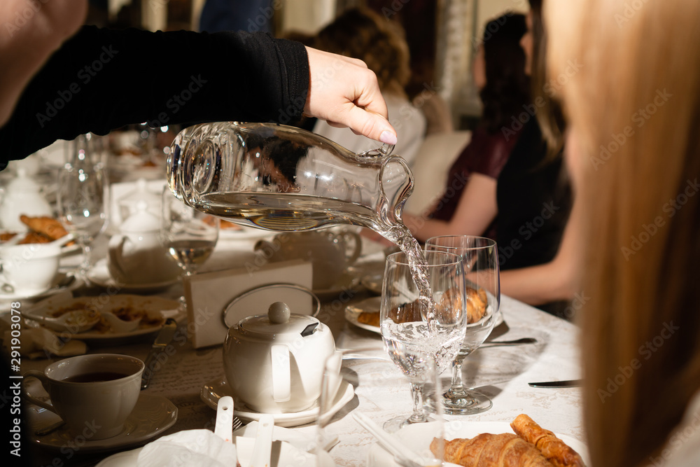 The gauntlet in black clothes holds transparent donut in water. Water pours into the pot. Covered table with white cups and white tablecloth. In the background are croissants. Horizontal orientation.