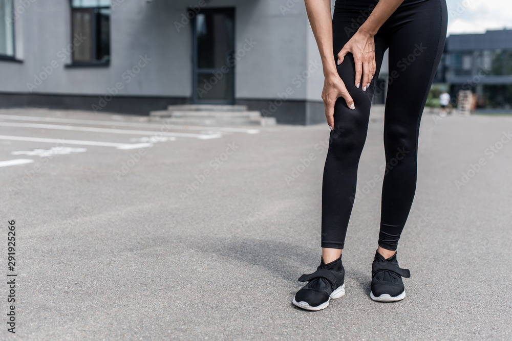 partial view of sportswoman in black sneakers with leg pain on street partial view of sportswoman in black sneakers with leg pain on street
