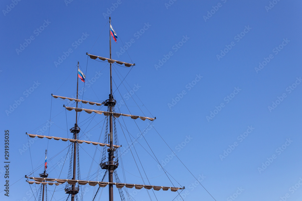 Mast of the ship with raised sails against the blue sky with flags